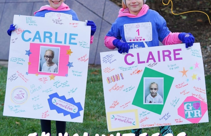 Two girls bundled up for fall weather holding cheers signs on a grassy lawn in a park. Text says 'Volunteers Needed.'